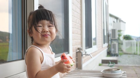 Asian toddler girl eats watermelon by both hands at backyard of house then smiling. A Japanese daughter in cute top knot hair enjoys summer season in the Mosquito coil smoke background