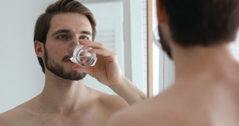 Smiling young healthy bearded guy holding glass, drinking pure fresh water, looking in mirror after showering. Happy handsome man enjoying daily healthcare habit, hydrating thirst in bathroom.