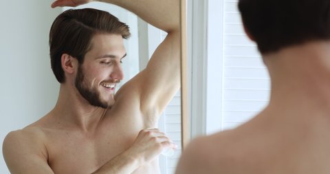 Handsome caucasian man looking in mirror, applying underarm deodorant in arm pit, preventing sweating. Happy young guy using antiperspirant after morning showering, enjoying daily skincare routine.