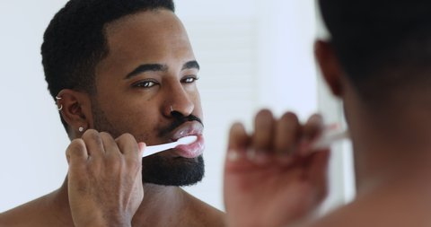 Close up head shot young handsome mixed race man brushing teeth in bathroom. Hipster african ethnicity guy looking in mirror, doing morning oral mouth hygienic daily procedure, toothcare concept.