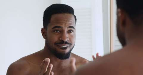 Mirror reflection head shot smiling charming young mixed race bare man self encouraging in bathroom. Happy shirtless african ethnicity guy doing confidence exercises, getting ready in morning.