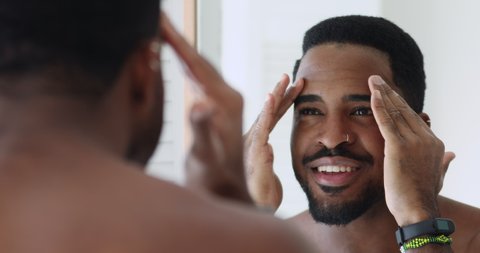 Head shot smiling african american hipster guy looks in mirror, touching facial skin, applying moisturizing product. Close up happy mixed race man using skincare products, satisfied with healthy look.