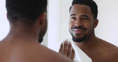 Happy young handsome shirtless afro ethnicity man looking in mirror, wiping wet face with white towel. Smiling mixed race hipster guy enjoying morning showering routine, getting ready in bathroom.