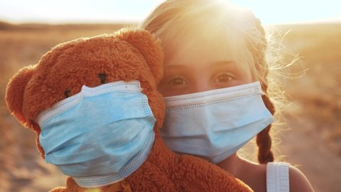 child girl in medical protective mask holding a teddy bear at sunset. concept pandemic coronavirus. girl kid face in a medical mask with covid -19 a teddy bear toy. girl during coronavirus quarantine