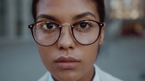 Close up face mixed race business woman in glasses standing look at camera serious outdoor in jacket posing at modern street success stylish exterior slow motion
