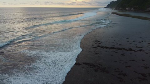 Wide shot of sandy ocean coastline at sunset. Small waves are sweeping the beach forming lot of white foam and licking the foots of dark cliffs on background. Aerial view, slow motion