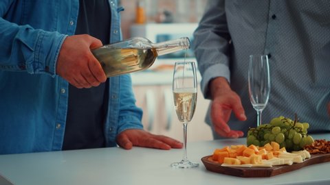 Close up of young man pouring white wine in glasses. Two generations tasting a cup of champagne in cozy dining room while women preparing the healthy dinner