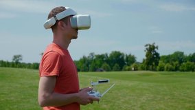 Man controls drone through remote control and looks video with goggles on head. Drone operator in a red T-shirt and blue shorts in park is filming video