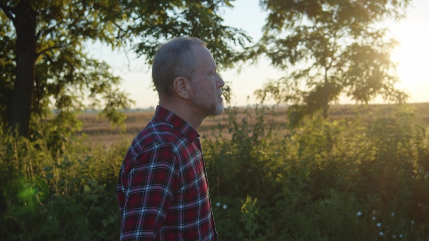 Side view of handsome bearded elderly man walking along the road enjoying beautiful sunset sunshine rising on background. Red casual shirt. Caucasian male person resting off the city urban life. 4K