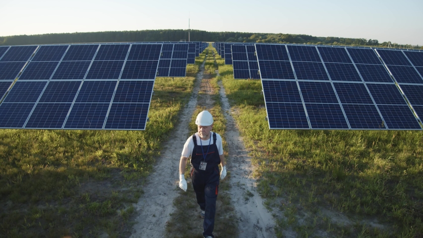 Professional mature man technician in hardhat walks on new ecological solar construction outdoors. Farm of solar panels. Concept of electricity, ecology, technologies. | Shutterstock HD Video #1057160278