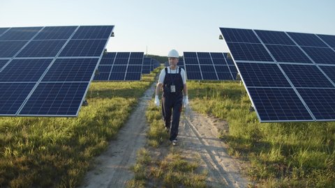 Professional mature man technician in hardhat walks on new ecological solar construction outdoors. Farm of solar panels. Concept of electricity, ecology, technologies.