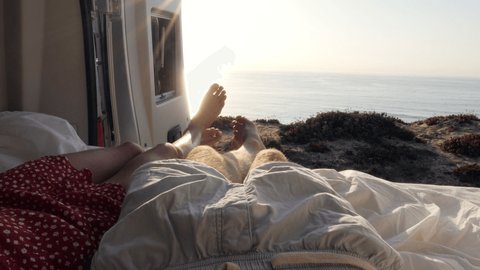 Point of view of couple's feet hanging out from van. Van lifestyle couple on camper van bed enjoys stunning ocean view 