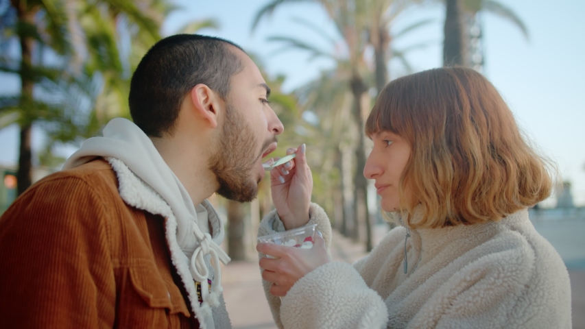 Pretty ginger caring female about lovely boyfriend feeding yogurt with spoon outdoor, happy young couple enjoying sunny day eating tasty dessert in plastic together travelers with palm trees Royalty-Free Stock Footage #1057169566