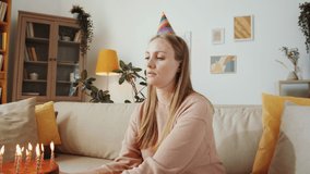 Beautiful blonde woman in party hat sitting on sofa at home, smiling and waving at camera, and then blowing candles on cake while celebrating birthday on video call during isolation