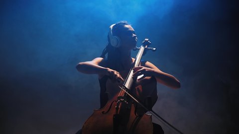 Young beautiful woman playing a melody on cello. Smoky dark studio