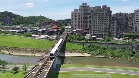 Aerial view of a metro train of Taipei MRT Wenhu Line traveling on a bridge over a riverside park and residential towers standing by the river on a sunny day in Taipei, the capital city of Taiwan