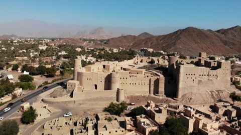 Bahla fort in Oman, famous touristic landmark surrounded by an arid landscape composed of sands, palms, medieval architecture, mountain ranges, under a blue sky