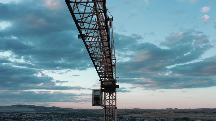 Tower crane against blue cloudy sky at the dusk and city horizon, industrial zone. Drone pan shot left to right on background field horizon, residential buildings