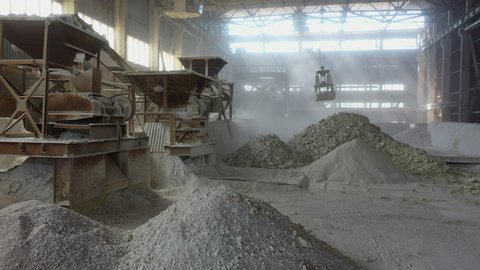 production workshop, crushing stones, huge bucket pick up stone blocks and lifts  into powerful unit. fractions and sorting on carpet line. rays of sun through dusty workshop, chemical industry grind
