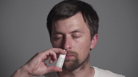 Young Caucasian man sprinkles medicine for rhinitis, colds and allergies into his nose from a white plastic bottle while sitting at home in the bedroom on the bed. Guy dripping nasal drops in nose.