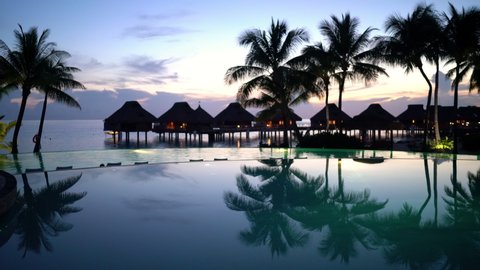 SEAMLESS LOOP VIDEO CINEMAGRAPH: Pool vacation travel destination - beach, palm trees and overwater bunglow hotel resort and luxury infinity swimming pool at amazing tropical sunset.
