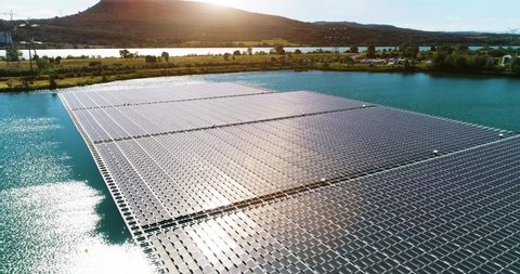 Floating solar panels in a blue pond under the sunlight. We can see the panels floating on water with mountains in the background - aerial view 4K