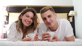 Portrait of young couple lying on bed holding cups of coffee and smiling at camera. Happy relationship. romantic scene in bedroom at home or hotel apartment. spending time in home interior. 4 k  - Powered by Shutterstock - Get 15% off with code: PIKWIZARD15