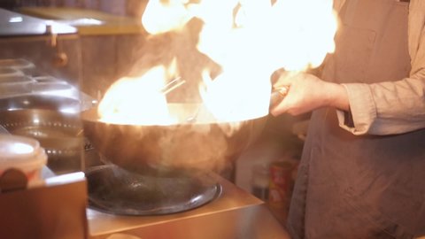 Chef cooking food with burning fire in slow motion. Close up shot of man hands and frying pan. Food in frying pan with flame on equipment kitchen background. Street food.
