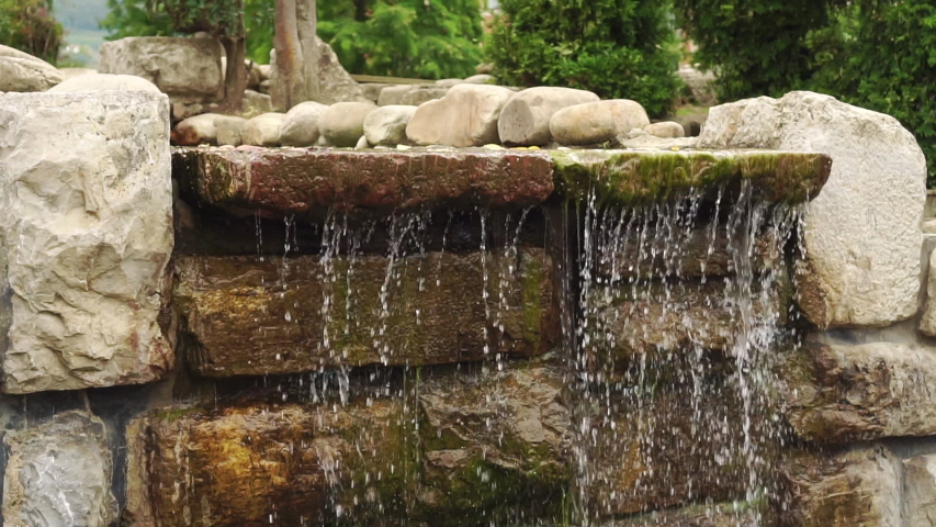 Waterfall of stone with moss in the park. The water flows down in streams and drops and falls down. Colorful summer landscape