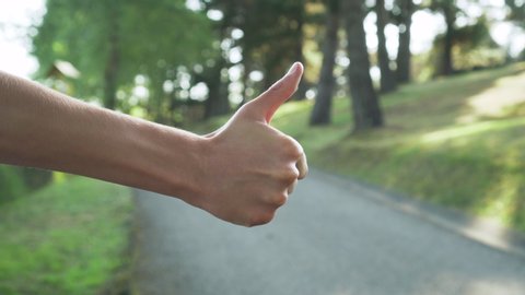 Hitchhiking close up. Hand with a raised male thumb on the background of the road and forest in blur. A tourist tries to stop the car on the road.