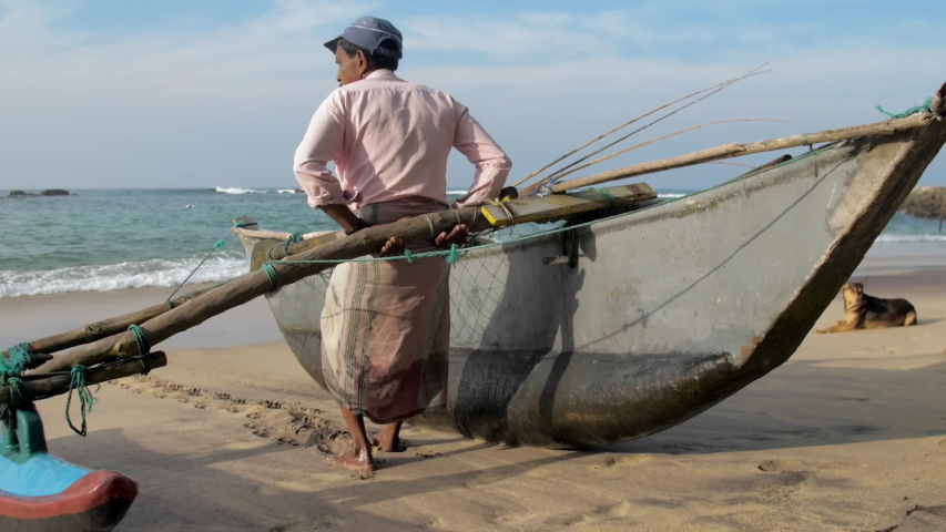 fisherman in long cloths holds and pulls traditional hand made fishing boat against sea close backside view slow motion. Concept Asian local people lifestyle