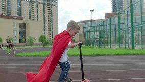 Slow motion video blond caucasian boy smiles cheerfully and rides scooter in red developing super hero cape, on sports field on sunny night. Boy has fun on walk riding skater