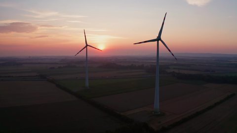 aerial photography drone shot of two wind turbines in the soft morning light, silhouettes in the early red dawn, sun rising at the horizon, highway with cars in the backgound
