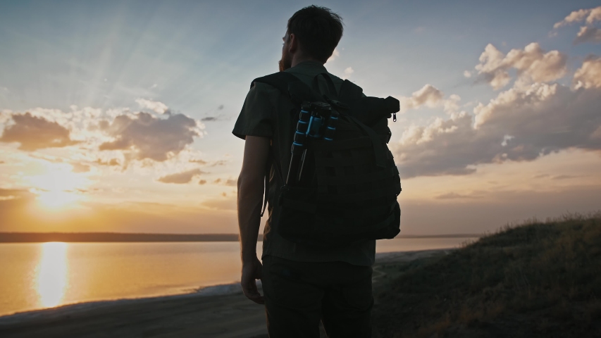 Young man photographer with backpack and tripod in it. He is standing on mountain, enjoying sunset and view on river.  