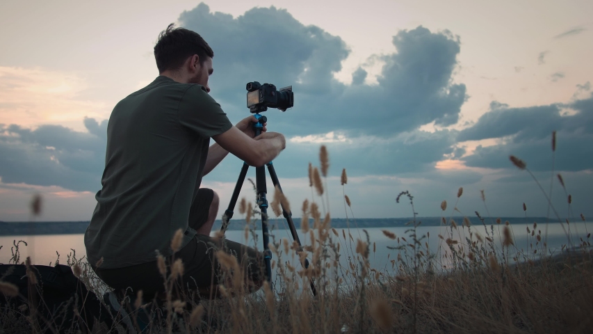 Male photographer sitting on high hill, adjusting camera fixed on tripod and making shoot. Picturesque view on huge lake. Summer evening, cloudy sky