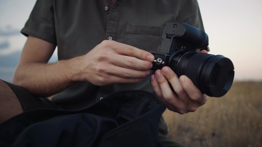 Unknown young man photographer with backpack is sitting on a meadow and preparing camera for fixation on tripod. Picturesque morning landscape