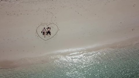 A drone shot of a couple lying on a pink sand beach in a heart drawn on the sand a small island near Maumere, Indonesia. Happy and careless moments. Waves gently washing the shore. Romance and love