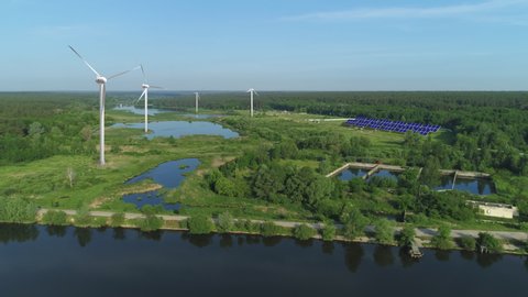 Aerial of wind turbines and solar panels on fish farm. Green trees and blue sky and water. Windmills and solar farm for energy production on beautiful blue sky. Wind power turbines and solar panels ge
