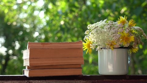 Closeup view 4k video footage of old paper books stacked on wooden background and cute sweet bunch of different wild meadow flowers arranged in blue metal vintage mug. Objects isolated at green blur.