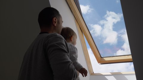 Father hold baby child and look through attic window, blue sky, fresh air, parent rising up child to discover nature, slow motion