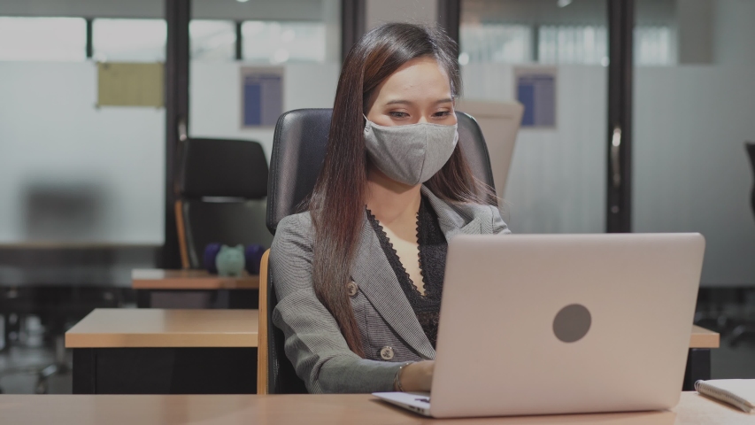 Medium shot: young Asian business woman working on computer and thinking of project in office. wearing protective face mask prevent corona virus after company reopen. new normal lifestyle concept | Shutterstock HD Video #1057232707