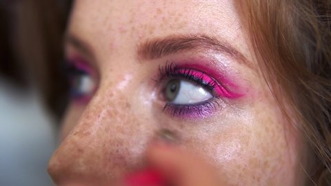 Unrecognizable makeup artist work in beauty studio. Woman applying tone using brush. Artist make a makeup for redhead model with freckles. Close up of eyes of a pretty girl with pink glitering make up
