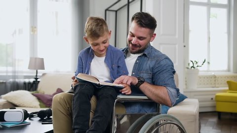 Likable satisfied positive young bearded man in wheelchair holding on knees his 8-aged son and helping him to read exciting book in contemporary living-room,slow motion