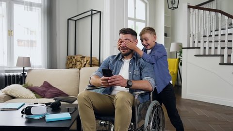 Handsome lucky cheerful small boy closing eyes of his surprised contented bearded father which sitting in wheelchair after accident and using smartphone