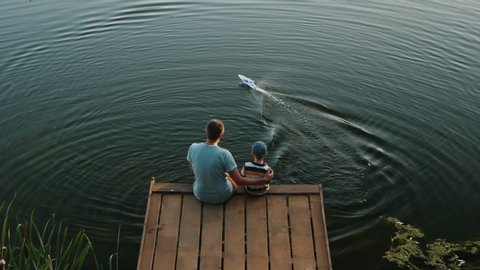Top view from the back of a dad with a little son sitting on a wooden pier playing with a RC speed-boat on the lake