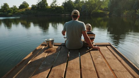 Rear view of a father with his little son are fishing while sitting on a wooden pier by the pond