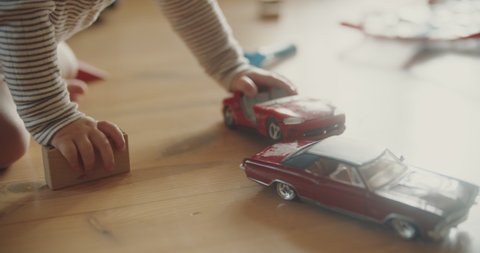 View of childs hands playing red car models and wooden blocks on the floor. Room filled with morning sunlight. Tiny fingers with toys.