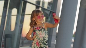 Kid girl at sunset wearing sunglasses uses her mobile phone in airport. Tourist child in overalls with flowers. Hawaiian style look. Using smartphone for video call, selfie. Vacations, tourism, trip