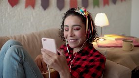 Lovely  brazilian woman dressed in red plaid shirt sitting in couch and watching live music concert on cellphone. Indoors at home living room. June Party holiday in Brazil, happiness, festival concept