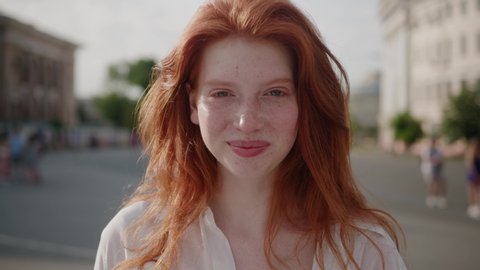 Face young sunshine woman with red hair look at camera smile stand in the city streets summer beautiful lady portrait happy outdoor close up slow motion